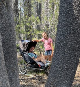 two girls in forest. one is in a wheelchair/stroller and the other is leaning on her chair